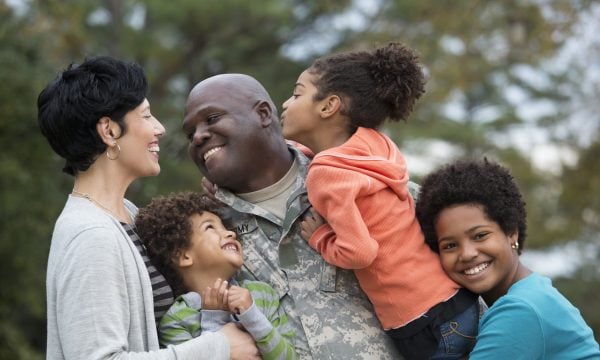 A father in army fatigues hugs his three children while smiling at his wife