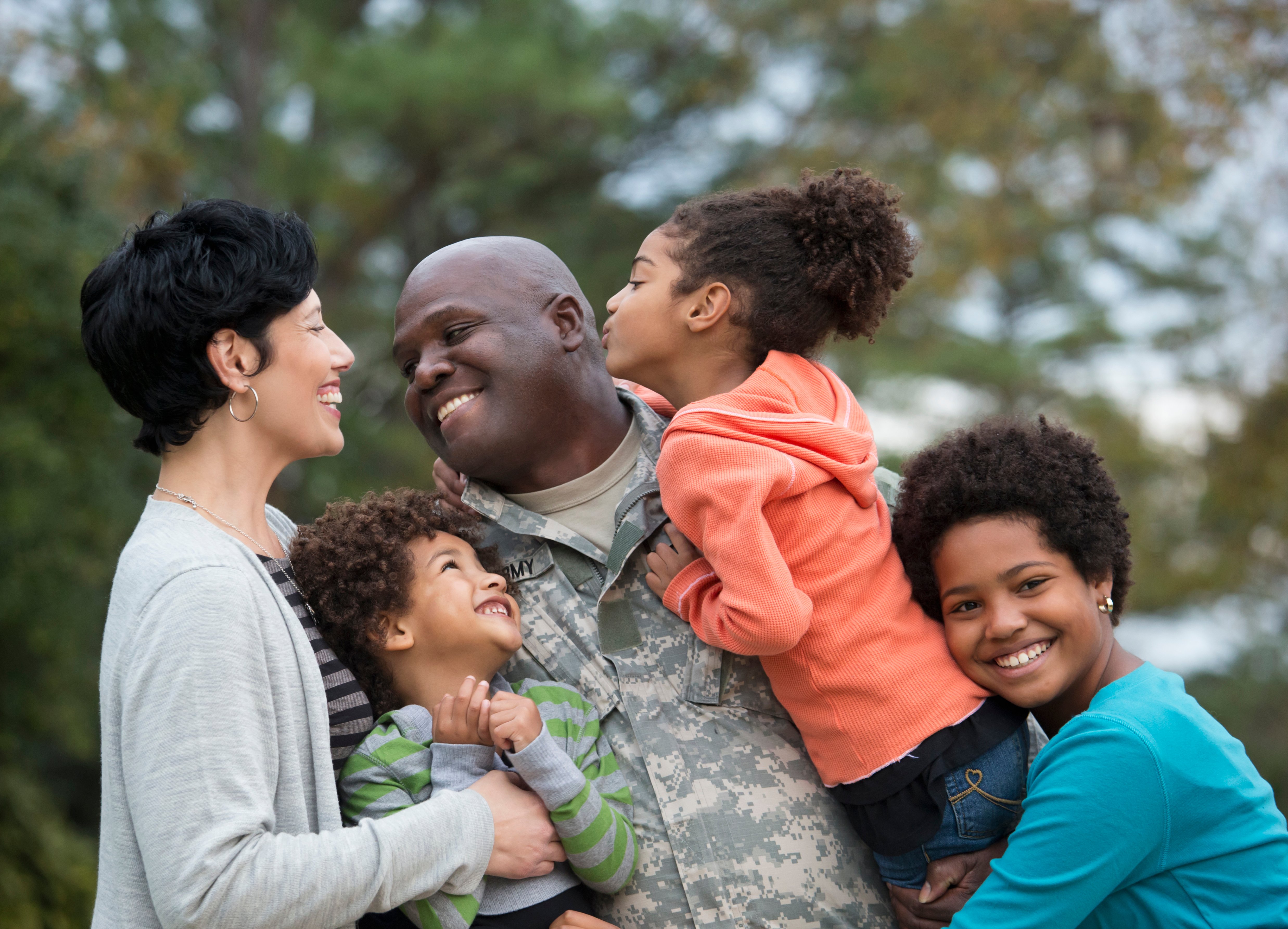 A father in army fatigues hugs his three children while smiling at his wife