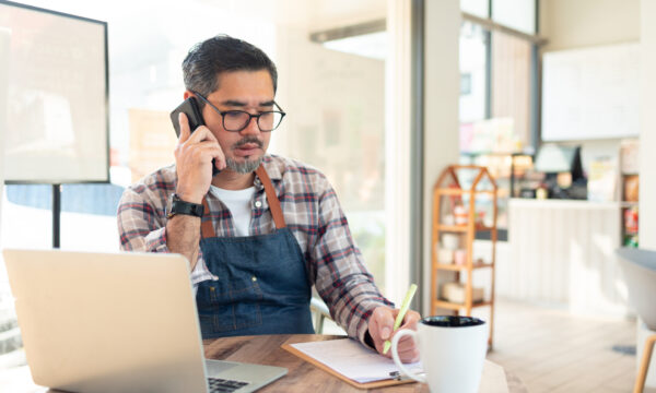 Entrepreneur talking on mobile phone while taking notes in front of his laptop. He sits in a modern coffee shop.