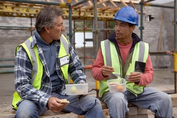 Two construction workers sitting down on a residential building site while eating their lunch.