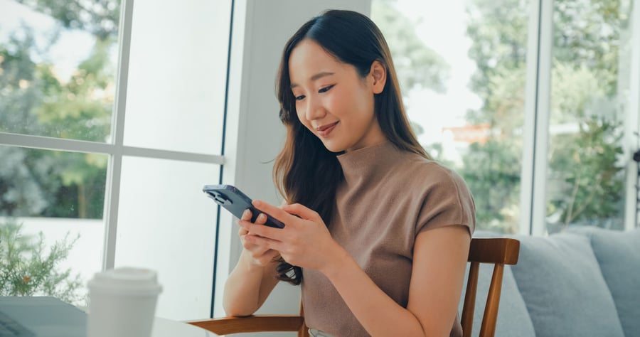 A young Asian woman is sitting in a cafe, using her smartphone to bank online.