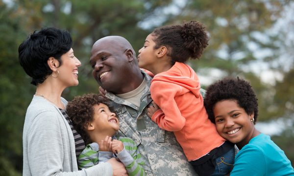 A father in army fatigues hugs his three children while smiling at his wife