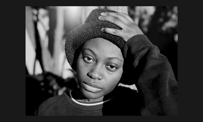 Funto looks directly into the camera, one hand holding up the brim of her hat, with trees behind.