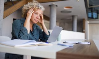 Professional woman suffering workplace stress, pressing fingers to temples while working on laptop in contemporary corporate setting